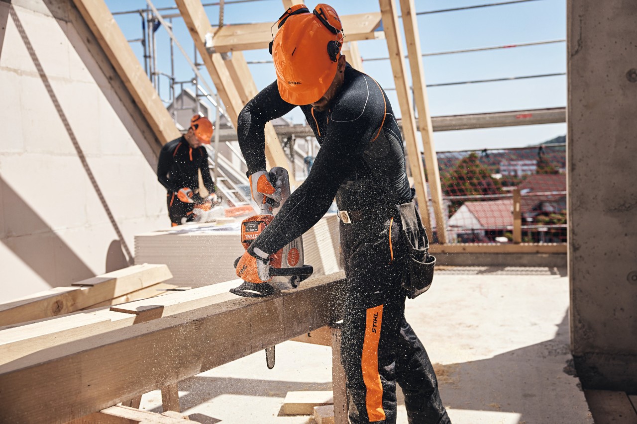 Ένας επαγγελματίας της STIHL σε έναν σκελετό στέγης τεμαχίζει μια ξύλινη δοκό με ένα επαναφορτιζόμενο αλυσοπρίονο, στο φόντο ένας ακόμα τεχνίτης εργάζεται με ένα επαναφορτιζόμενο αλυσοπρίονο